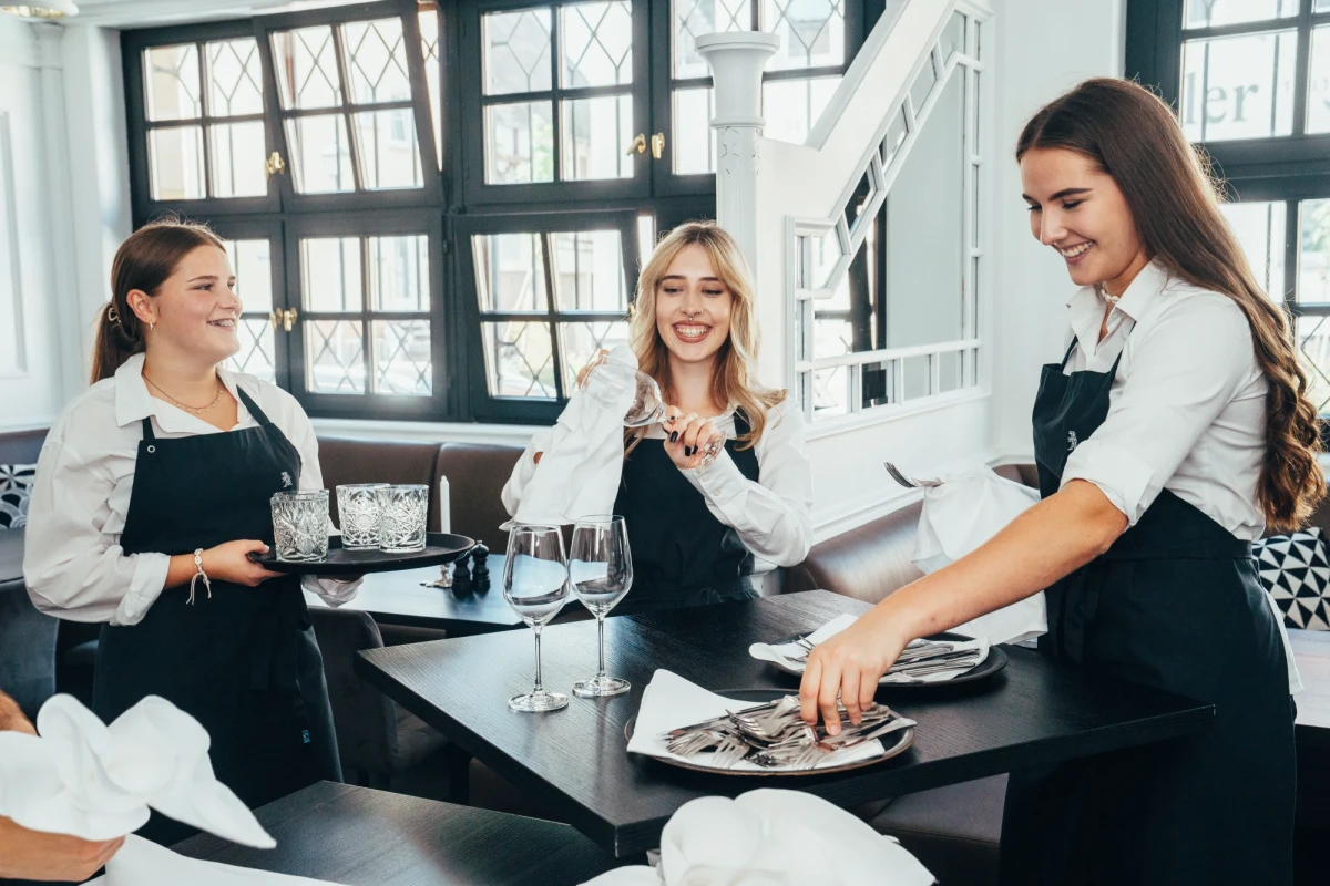 Drei Kellnerinnen in schwarzen Sch&uuml;rzen bereiten einen Tisch in einem hellen, modernen Restaurant vor. Sie l&auml;cheln und arbeiten gemeinsam mit Tellern und Weingl&auml;sern. Die gro&szlig;e Fensterfront im Hintergrund l&auml;sst viel nat&uuml;rliches Licht in den Raum.