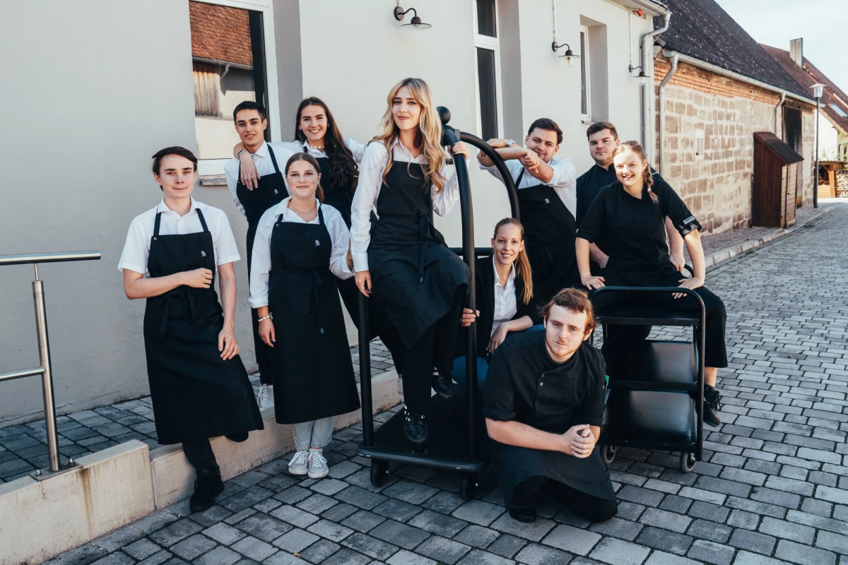 Eine Gruppe von neun Mitarbeitern in schwarzen Sch&uuml;rzen steht und sitzt auf einer gepflasterten Stra&szlig;e vor einem Geb&auml;ude, sie posieren als Team in einer lockeren, freundlichen Aufstellung.