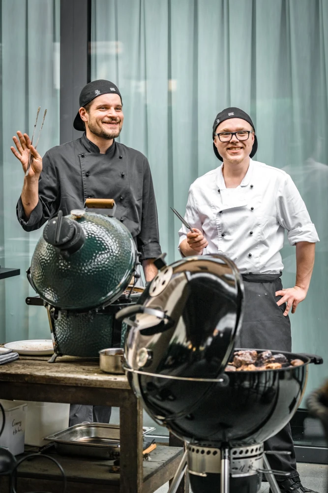 Zwei K&ouml;che in Berufskleidung stehen neben gro&szlig;en Grillger&auml;ten, einer in grauer und einer in wei&szlig;er Jacke, beide l&auml;chelnd und in einer entspannten Arbeitshaltung.