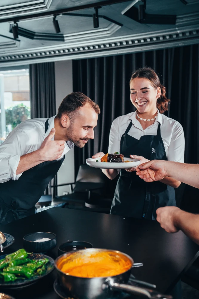 Ein K&uuml;chenchef in schwarzer Sch&uuml;rze und eine l&auml;chelnde Servicekraft in wei&szlig;em Hemd und schwarzer Sch&uuml;rze bereiten gemeinsam ein Gericht in einer professionellen Restaurantk&uuml;che zu.