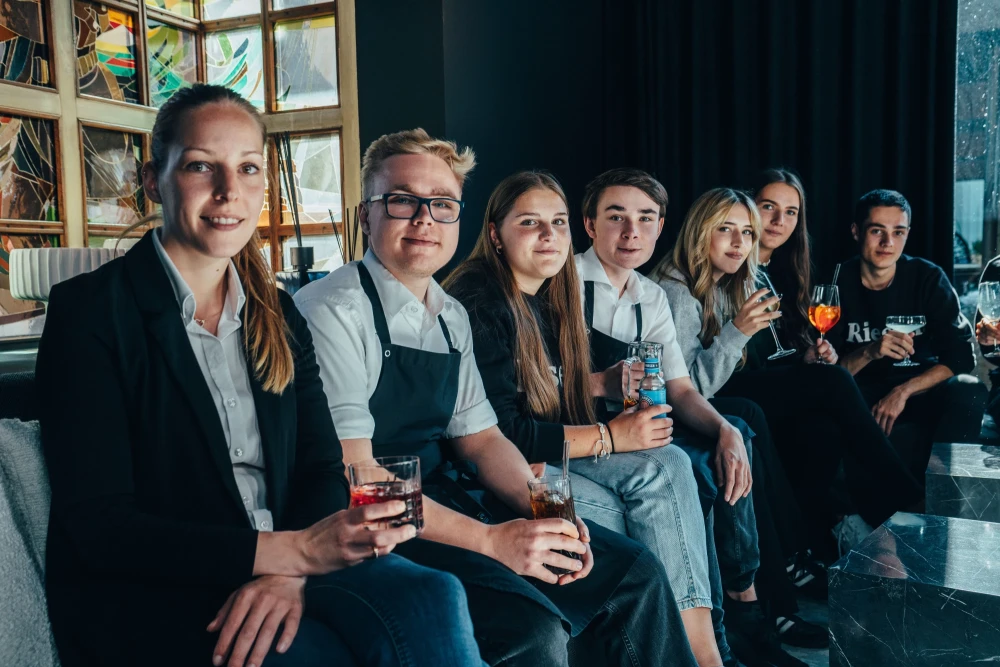 Sieben Mitarbeitende des Hotel Riesengebirge sitzen mit gef&uuml;llten Getr&auml;nkegl&auml;sern in der Hand auf einem Sofa in der Bar des Hotel Riesengebirge. Alle blicken freundlich in die Kamera