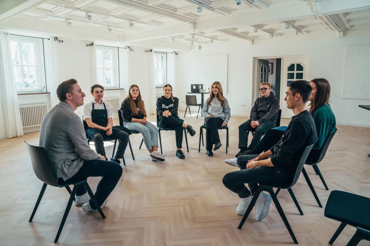 Gruppe von jungen Menschen sitzt in einem Stuhlkreis in einem Tagungsraum des Hotel Riesengebirge mit dem Hoteldirektor. Es herrscht eine lockere Stimmung.
