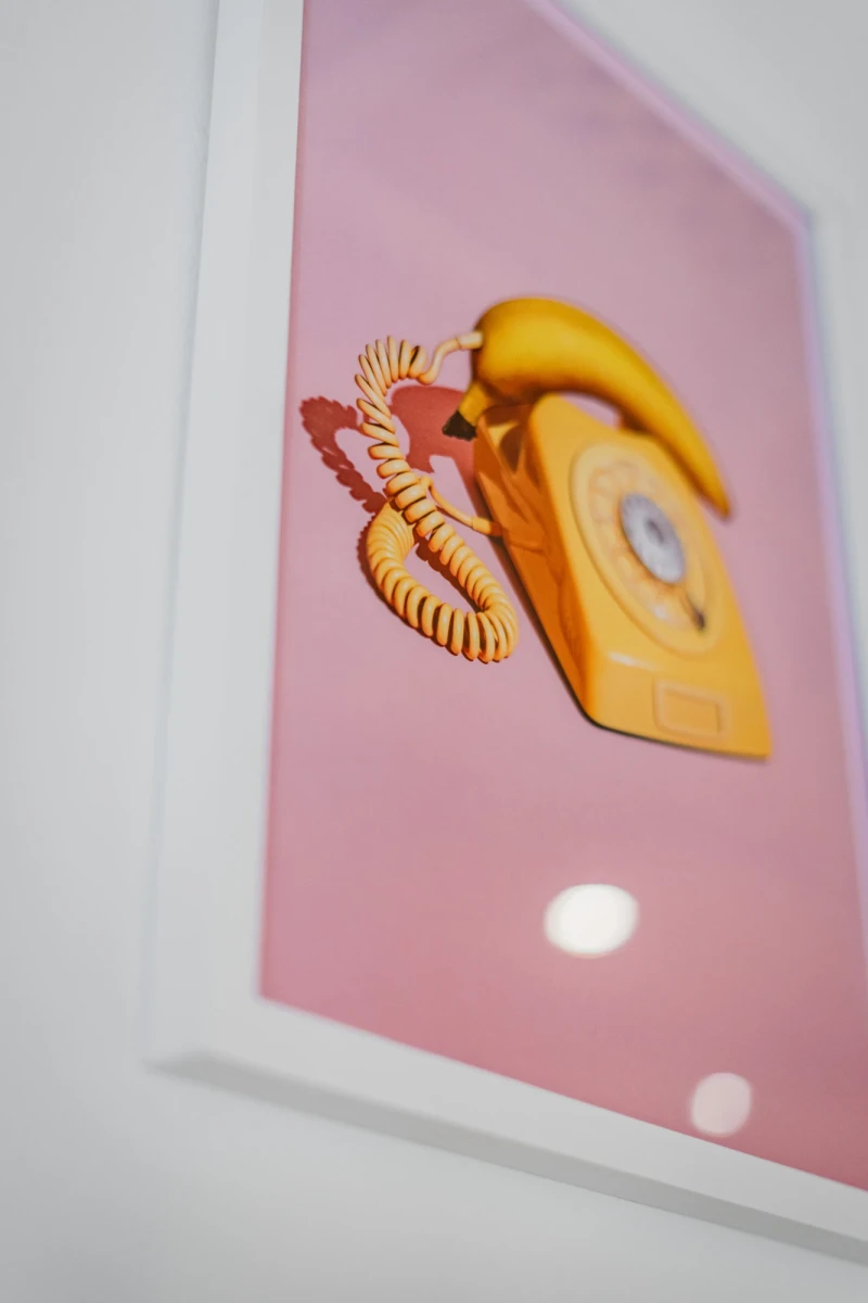 Gelbes Vintage-Telefon mit gedrehter Schnur auf einem rosa Hintergrund, das leicht schr&auml;g in einem wei&szlig;en Rahmen positioniert ist und eine nostalgische, minimalistisch gestaltete Atmosph&auml;re ausstrahlt.