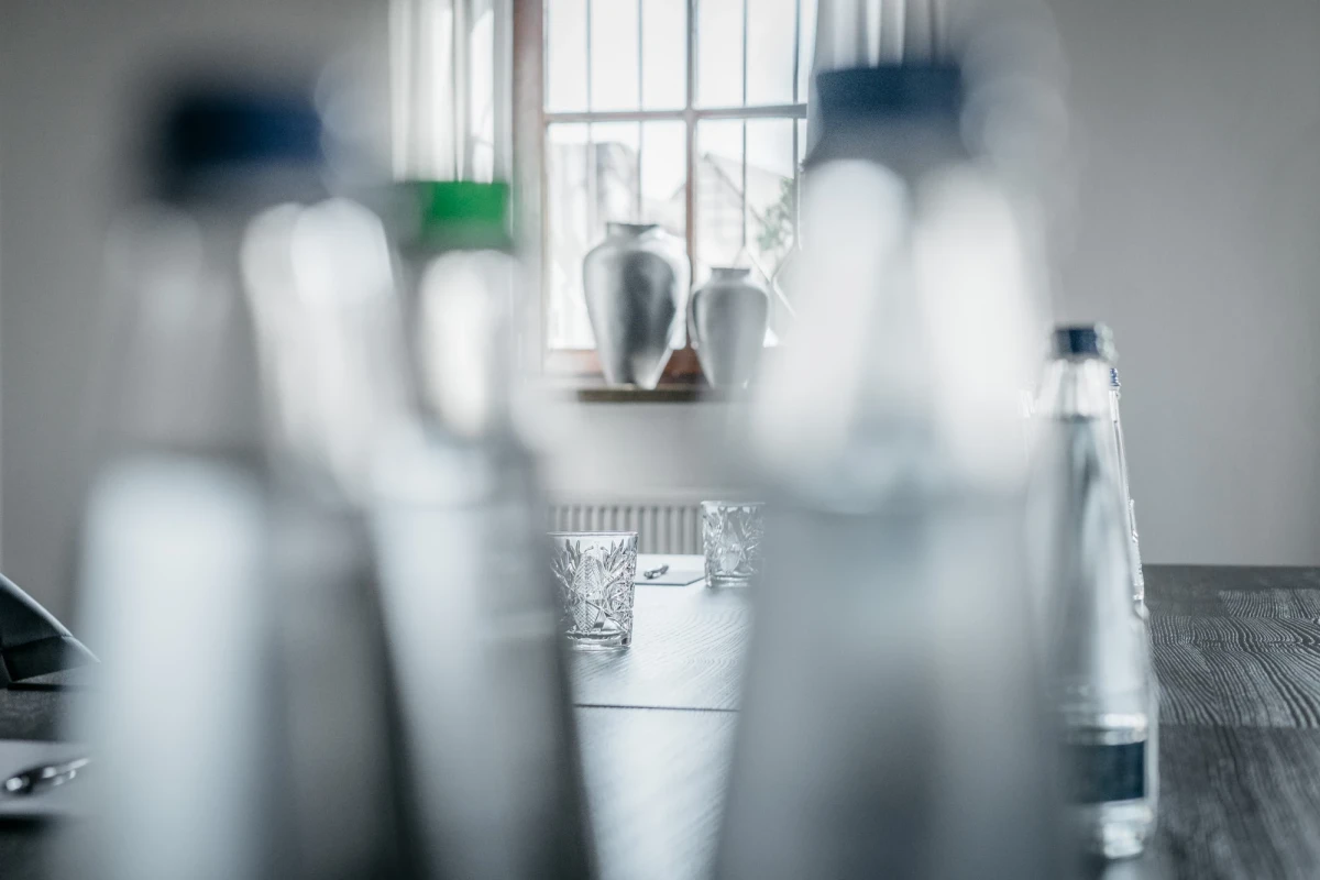 Mehrere unscharfe Glasflaschen im Vordergrund, im Hintergrund ein Fenster mit Vorh&auml;ngen und einem Blumenstrauss. Ein Kristallglas steht auf einem Holztisch, der Raum wirkt ged&auml;mpft und minimalistisch.