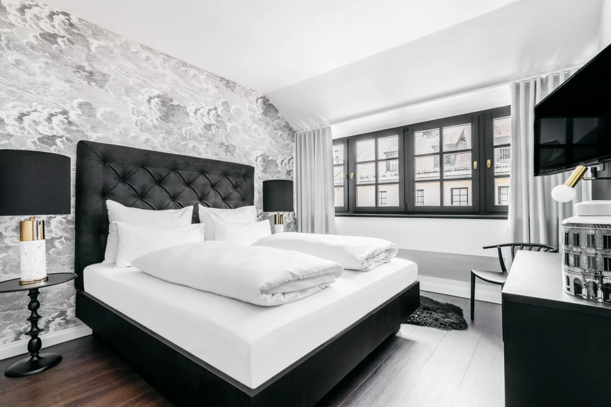 Modernes Hotelzimmer in Schwarz-Wei&szlig; mit gepolstertem Bett, gemusteter Akzentwand und gro&szlig;em Fenster. Dunkler Holzboden, wei&szlig;e Bettw&auml;sche und elegante schwarze Nachttischlampen komplettieren das stilvolle Interieur.
