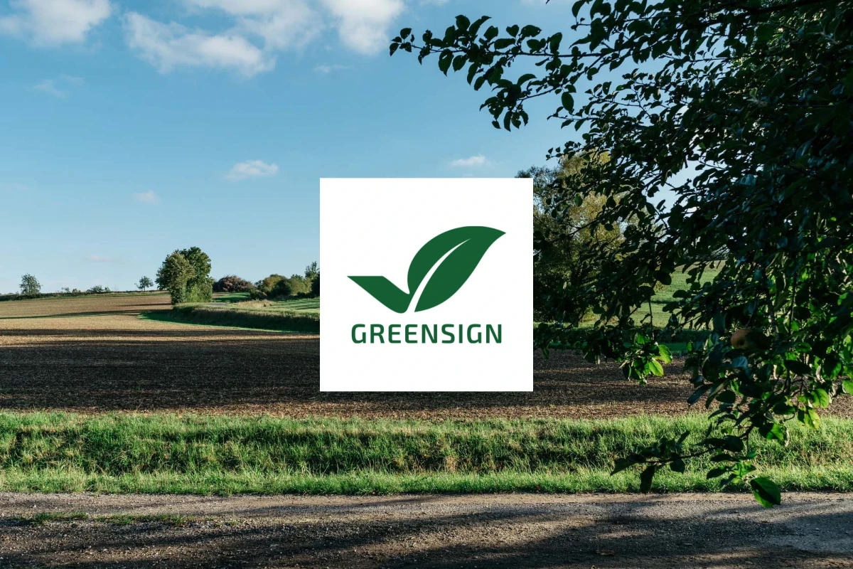 Gr&uuml;ne Landschaft mit Feldern, B&auml;umen und einem wei&szlig;en Quadrat mit dem Greensign-Logo in der Mitte. Das Logo zeigt ein stilisiertes gr&uuml;nes Blatt mit einem H&auml;kchen.