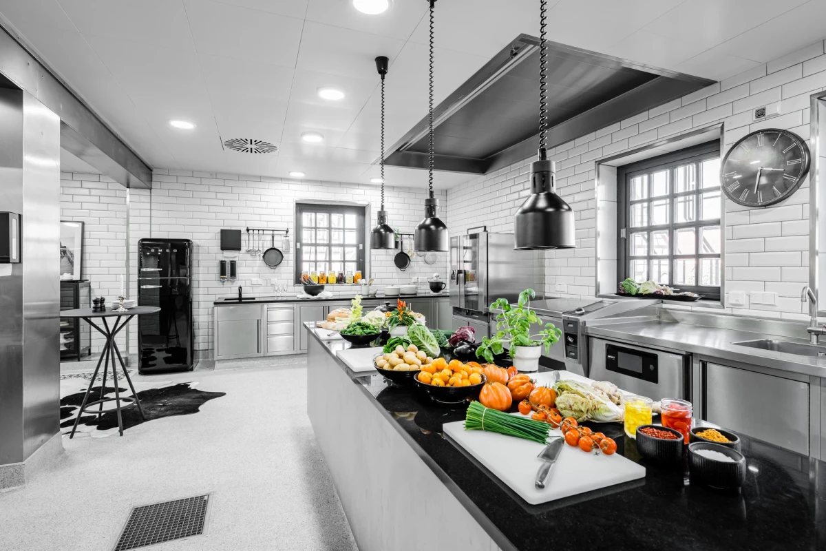 Moderne, ger&auml;umige Profik&uuml;che mit wei&szlig;en Kacheln und schwarzer Arbeitsplatte, auf der frisches Gem&uuml;se und Obst wie Orangen, Paprika und gr&uuml;ne Blattgem&uuml;se ausgelegt sind. Helle Industriebeleuchtung und Edelstahlger&auml;te pr&auml;gen die professionelle Atmosph&auml;re der K&uuml;che.