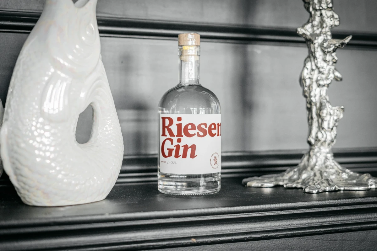 Flasche Rieser Gin auf einem dunklen Regal zwischen einer wei&szlig;en Keramikvase und einem silbernen Kerzenhalter. Die Flasche hat ein klares Design mit rotem Schriftzug und Korken.