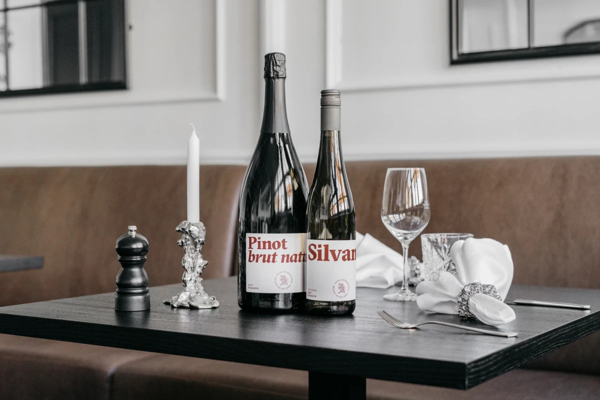 Zwei Weinflaschen von Pinot Brut nah und Silvar stehen auf einem dunklen Holztisch neben einem Weinglas, einer Kerze und einer Pfefferm&uuml;hle in ged&auml;mpften Farbt&ouml;nen.