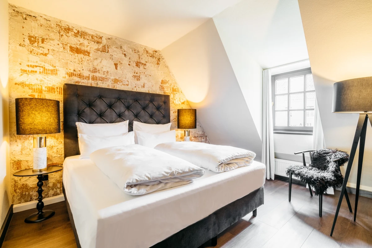 Ger&auml;umiges Hotelzimmer mit gro&szlig;em gepolstertem Kopfteil und wei&szlig;er Bettw&auml;sche. Warme Beleuchtung und eine Akzentwand mit vintage Tapete, daneben ein Fenster mit Tageslicht. Ein gemusterter Sessel und eine Stehlampe vervollst&auml;ndigen die einladende Atmosph&auml;re.