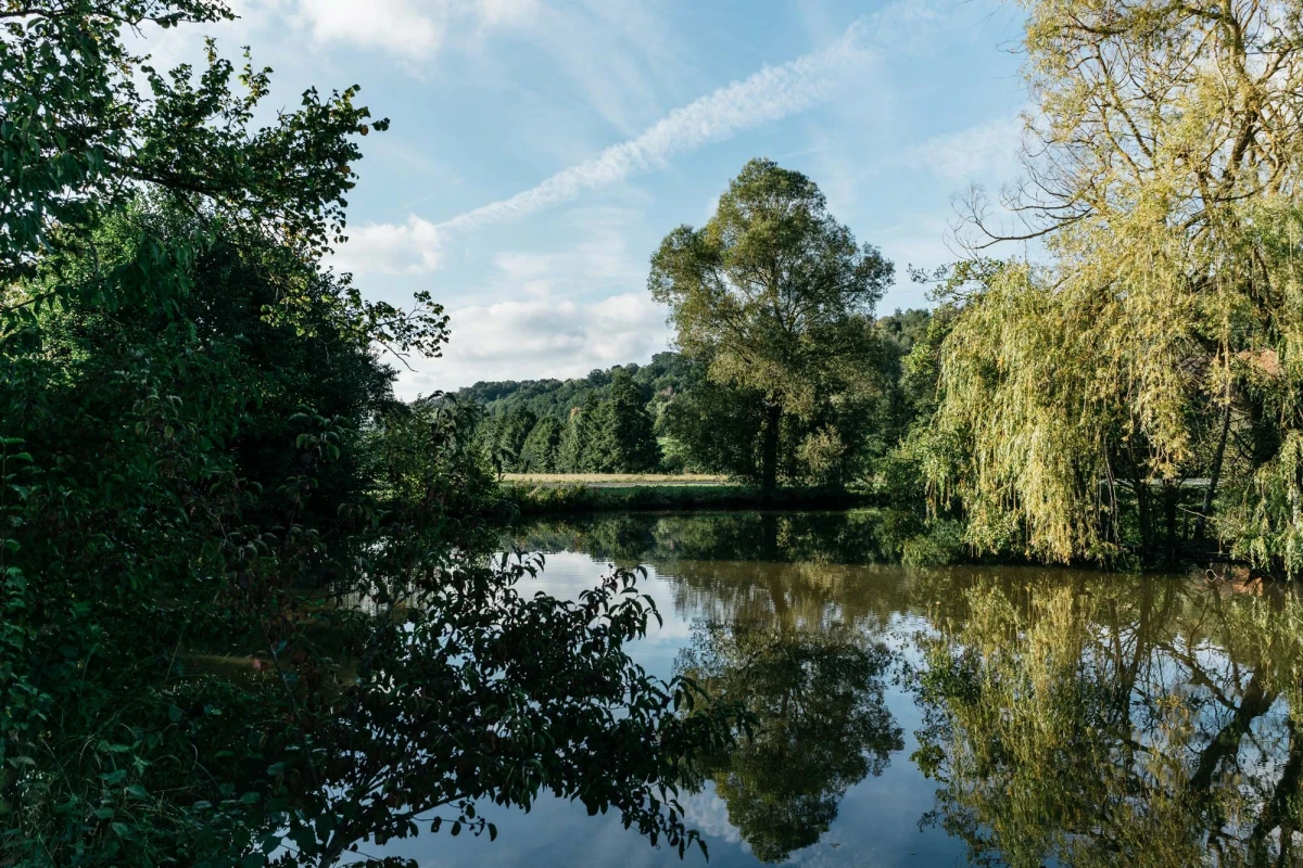 Ruhige Teichlandschaft mit spiegelblanker Wasseroberfl&auml;che, umgeben von dicht bewachsenen Ufern mit Laubb&auml;umen in verschiedenen Gr&uuml;nt&ouml;nen. Wei&szlig;liche Schleierwolken am blauen Himmel rahmen die friedvolle Naturszene ein.