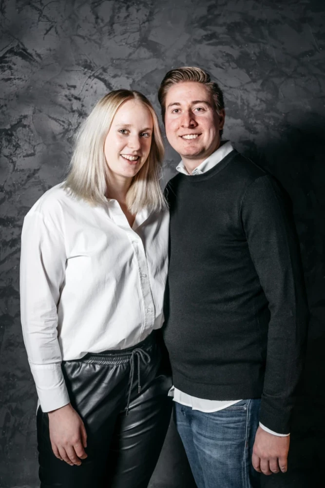 Zwei l&auml;chelnde Personen vor einer grauen Betonwand. Eine Frau in wei&szlig;er Bluse und Lederhose steht links, ein Mann in schwarzem Pullover und blauer Jeans rechts von ihr. Beide stehen eng beieinander und wirken freundlich.