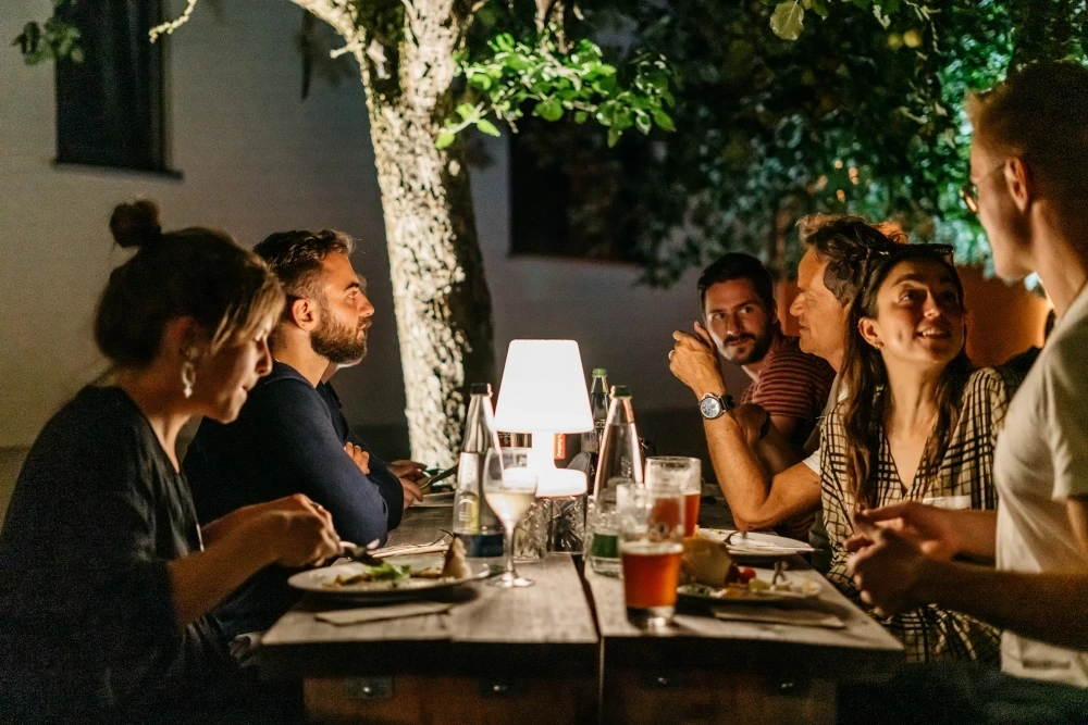 Mehrere Personen sitzen abends an einem Tisch im Freien, beleuchtet von einer Tischlampe. Sie unterhalten sich und essen gemeinsam, w&auml;hrend im Hintergrund ein beleuchteter Baum steht.
