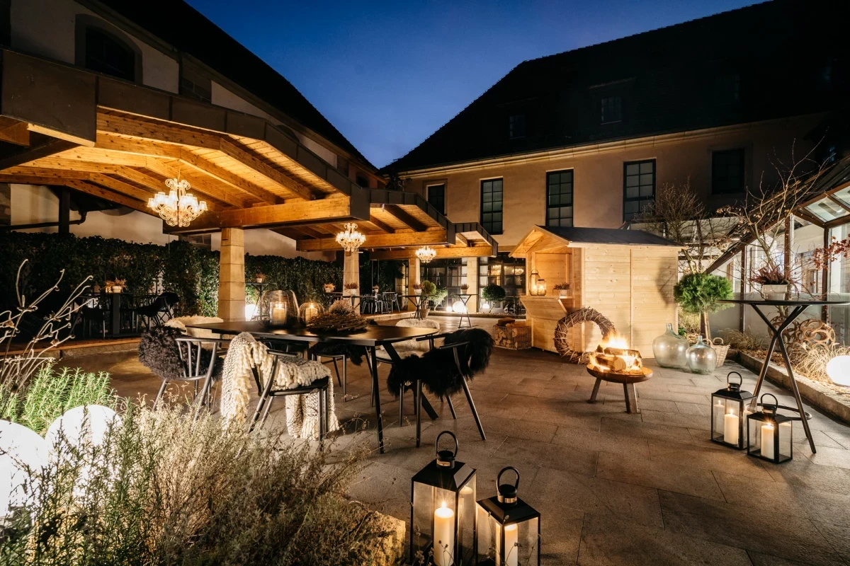 Abendliche Terrasse mit &uuml;berdachtem Sitzbereich, warm beleuchtet von Laternen und H&auml;ngelampen. Gem&uuml;tliche Atmosph&auml;re mit Gartenm&ouml;beln, Feuerstelle und dekorativen Laternen vor einer Hausfassade bei Nacht.