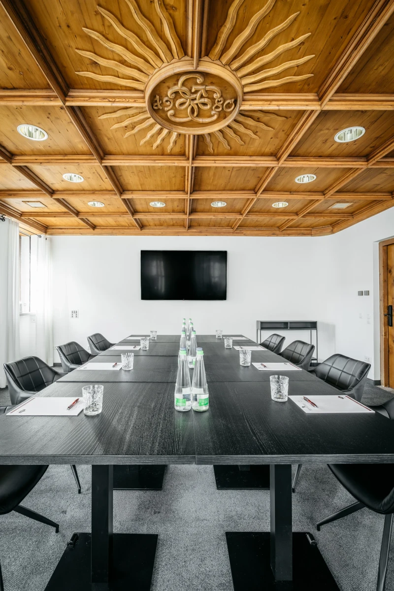 Moderner Konferenzraum mit schwarzem Konferenztisch, grauen Lederst&uuml;hlen und einer Holzbalkendecke mit einem sonnenstrahlenf&ouml;rmigen Zentralmotiv. An der Wand h&auml;ngt ein gro&szlig;er schwarzer Bildschirm, und auf dem Tisch stehen Wassergl&auml;ser und Notizbl&ouml;cke f&uuml;r Besprechungen.