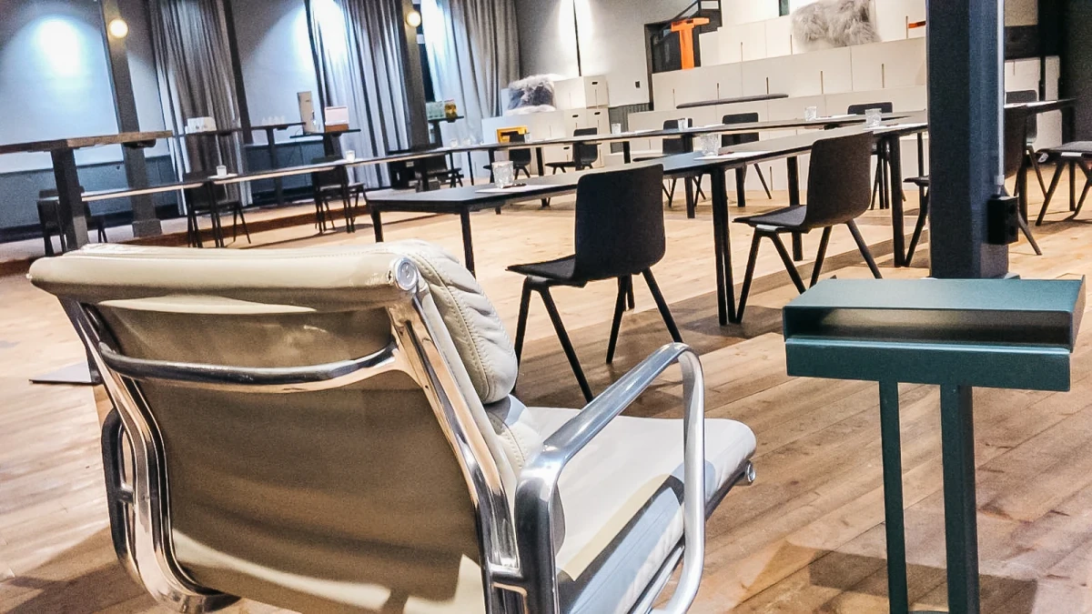 Moderner Seminarraum mit holzfarbenen B&ouml;den, schwarzen St&uuml;hlen und einer wei&szlig;en Sitzgruppe im Vordergrund. Lichtdurchfluteter Raum mit Gardinen und minimalistischer Einrichtung, der auf Professionalit&auml;t und Funktionalit&auml;t ausgelegt ist.