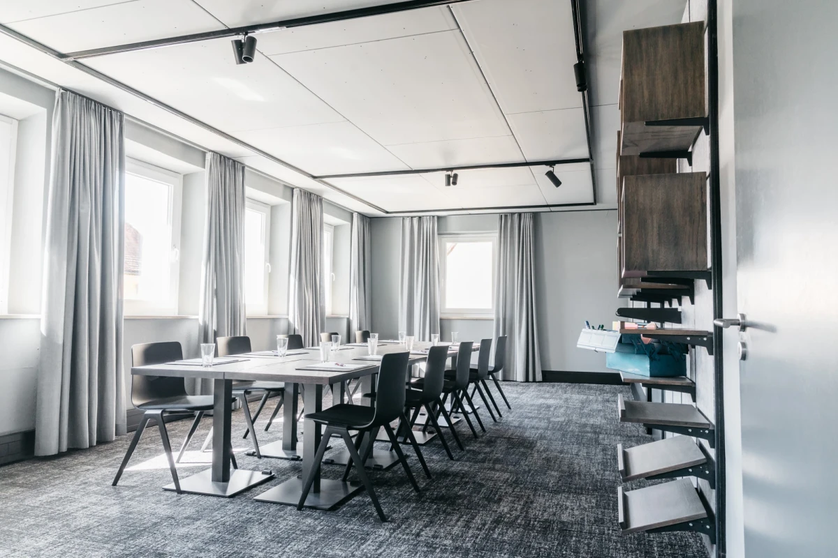 Moderner Konferenzraum mit gro&szlig;en Fenstern und grauen Vorh&auml;ngen. Lange wei&szlig;e Tische mit schwarzen St&uuml;hlen sind auf einem grauen Teppich angeordnet. An der Seite befindet sich ein Holzregal mit Dokumentenablagen.