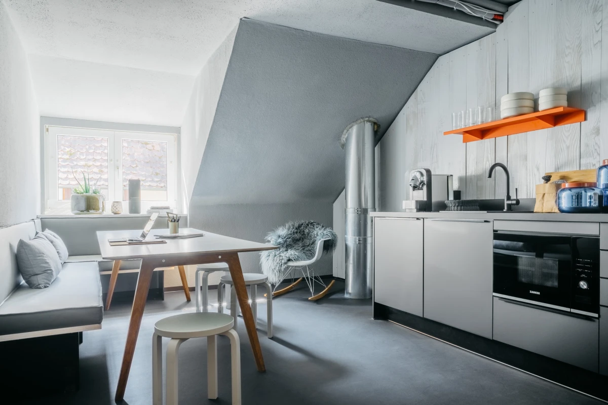 Moderne Dachgeschoss-K&uuml;che mit wei&szlig;en Schr&auml;nken, einem orangefarbenen Regal und einem hellen Holztisch mit St&uuml;hlen. Graue W&auml;nde, bodentiefe Fenster und eine minimalistische Einrichtung pr&auml;gen den zeitgen&ouml;ssischen Wohnraum.