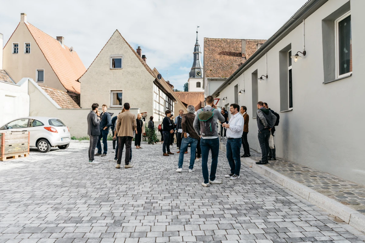Eine Gruppe von Menschen steht auf einem gepflasterten Platz mit historischen Geb&auml;uden und einem Kirchturm im Hintergrund. Die Teilnehmer sind in Gr&uuml;ppchen verteilt und tragen legere Kleidung, w&auml;hrend sie sich offenbar im Gespr&auml;ch austauschen.