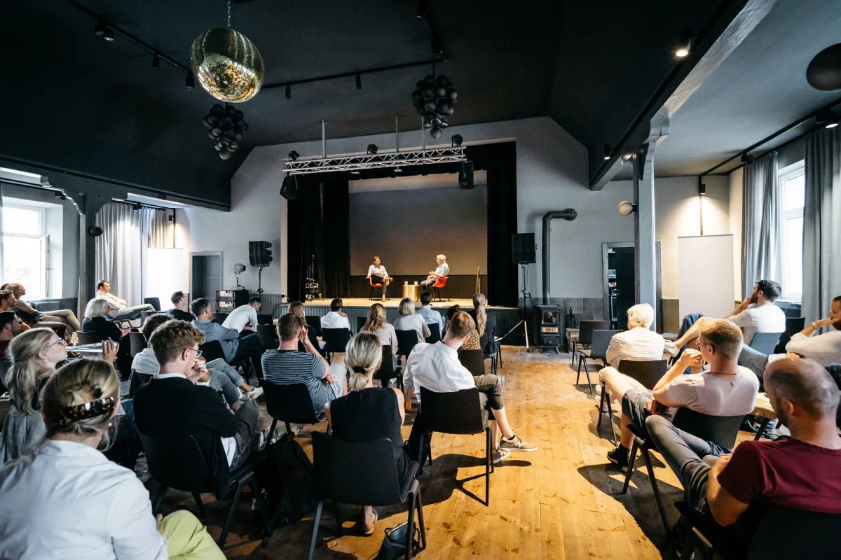 Zwei Personen sitzen auf einer B&uuml;hne vor einem Publikum, das aufmerksam einer Pr&auml;sentation oder Diskussion in einem hell erleuchteten Veranstaltungsraum mit Holzboden und Discokugel folgt.