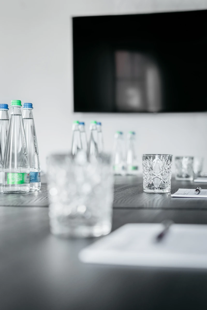 Glasflaschen mit Wasser und dekorativ geschliffene Wassergl&auml;ser auf einem dunklen Tisch mit unscharfem Hintergrund, der einen Teil eines schwarzen Bildschirms oder Fernsehers zeigt.