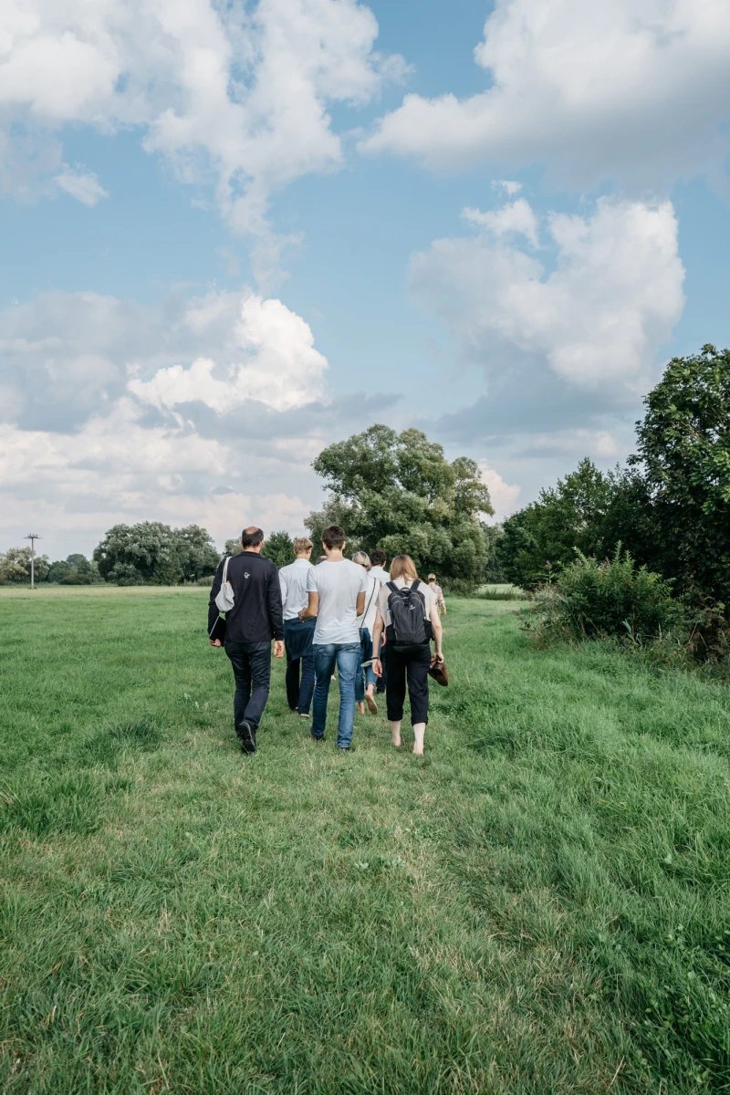 F&uuml;nf Personen gehen eine gr&uuml;ne Wiese entlang, im Hintergrund B&auml;ume und ein blauer Sommerhimmel mit wei&szlig;en Wolken. Die Gruppe tr&auml;gt Alltagskleidung und bewegt sich von hinten gesehen.