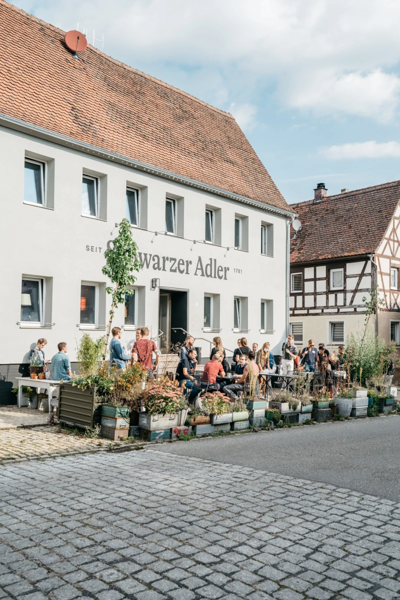 Stra&szlig;enszene vor dem Gasthaus "Schwarzer Adler" mit Fachwerkh&auml;usern, belebtem Au&szlig;enbereich und Besuchern, die an Tischen und Pflanzk&uuml;beln vor dem wei&szlig;en Geb&auml;ude sitzen.