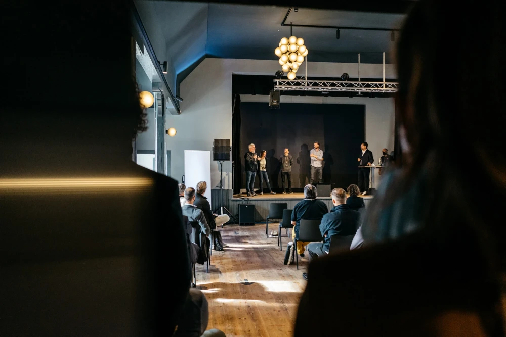 Mehrere Personen stehen auf einer B&uuml;hne vor einem Publikum in einem Veranstaltungsraum mit Holzboden und elegantem Kronleuchter. Die Atmosph&auml;re wirkt professionell und konzentriert, mit Zuh&ouml;rern, die der Pr&auml;sentation aufmerksam folgen.