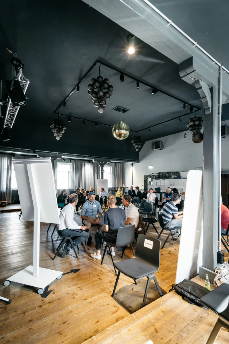 Seminarraum mit hoher Decke und Holzboden, in dem mehrere Personen an Tischen sitzen und an einem Workshop oder einer Konferenz teilnehmen. Moderne Beleuchtung und wei&szlig;e Stellw&auml;nde pr&auml;gen den hellen, industriell anmutenden Raum.