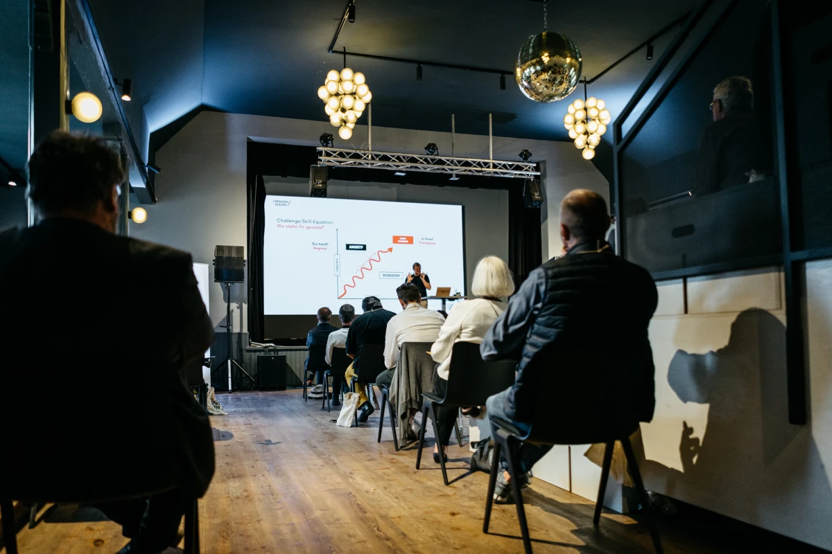 Pr&auml;sentator spricht vor Publikum in einem modern gestalteten Raum mit Holzboden und h&auml;ngenden Kristallleuchtern. Ein Projektionsbildschirm zeigt Pr&auml;sentationsfolien, w&auml;hrend Zuh&ouml;rer aufmerksam auf St&uuml;hlen sitzen.