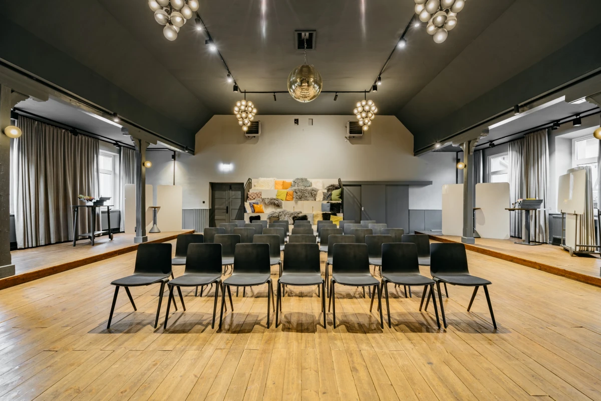 Moderner Konferenzraum mit h&ouml;lzernem Fu&szlig;boden, grauen W&auml;nden und schwarzen St&uuml;hlen, die in Reihen aufgestellt sind. Gro&szlig;e Deckenleuchten und Vorh&auml;nge an den Seitenw&auml;nden sorgen f&uuml;r eine einladende Atmosph&auml;re. Ein Kunstwerk in gelb-grauen T&ouml;nen schm&uuml;ckt die R&uuml;ckwand des Raumes.