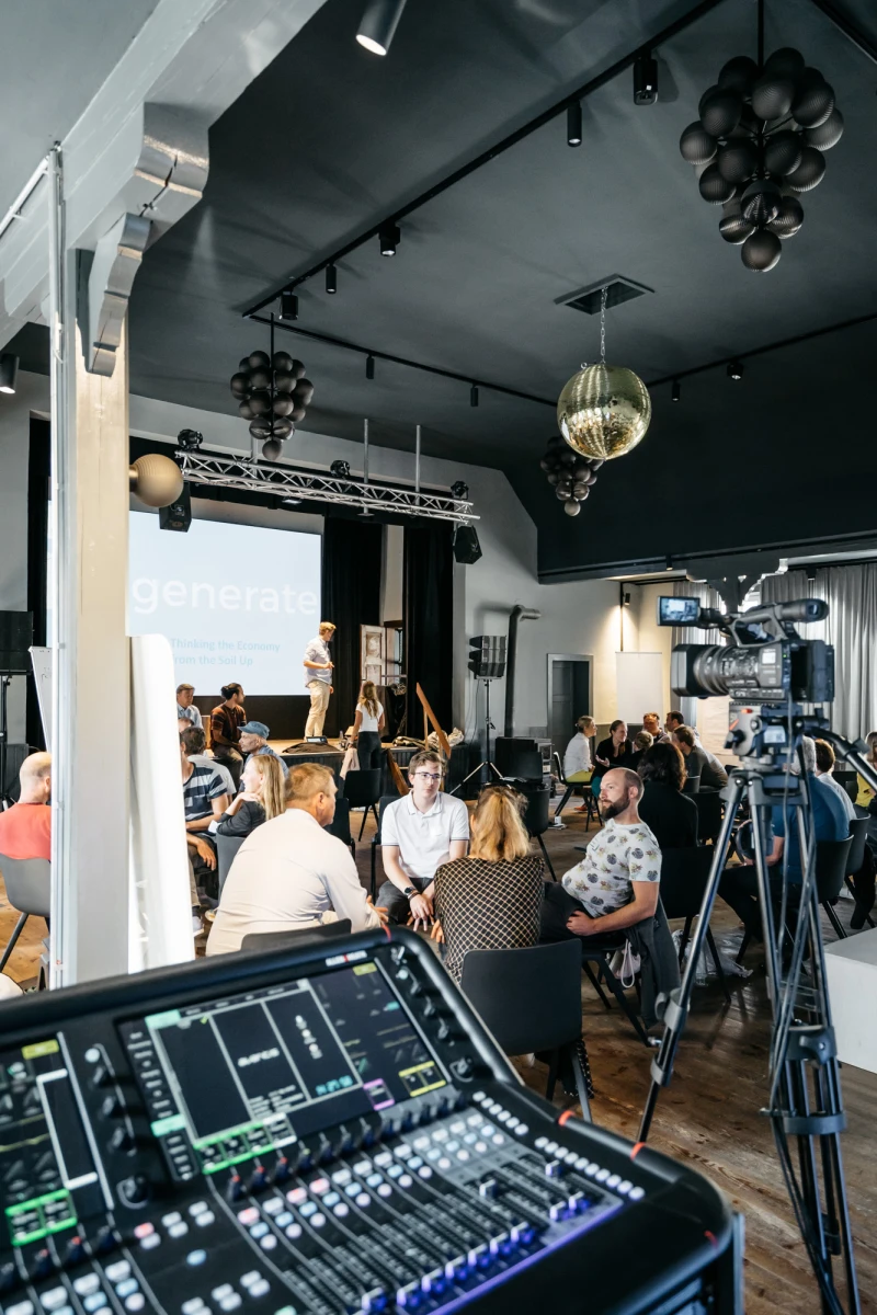 Audio-Produktionsraum mit Mischpult im Vordergrund, Kamera und mehreren Personen im Hintergrund, die an einem technischen Setup in einem modern eingerichteten Raum mit Deckenbeleuchtung arbeiten.