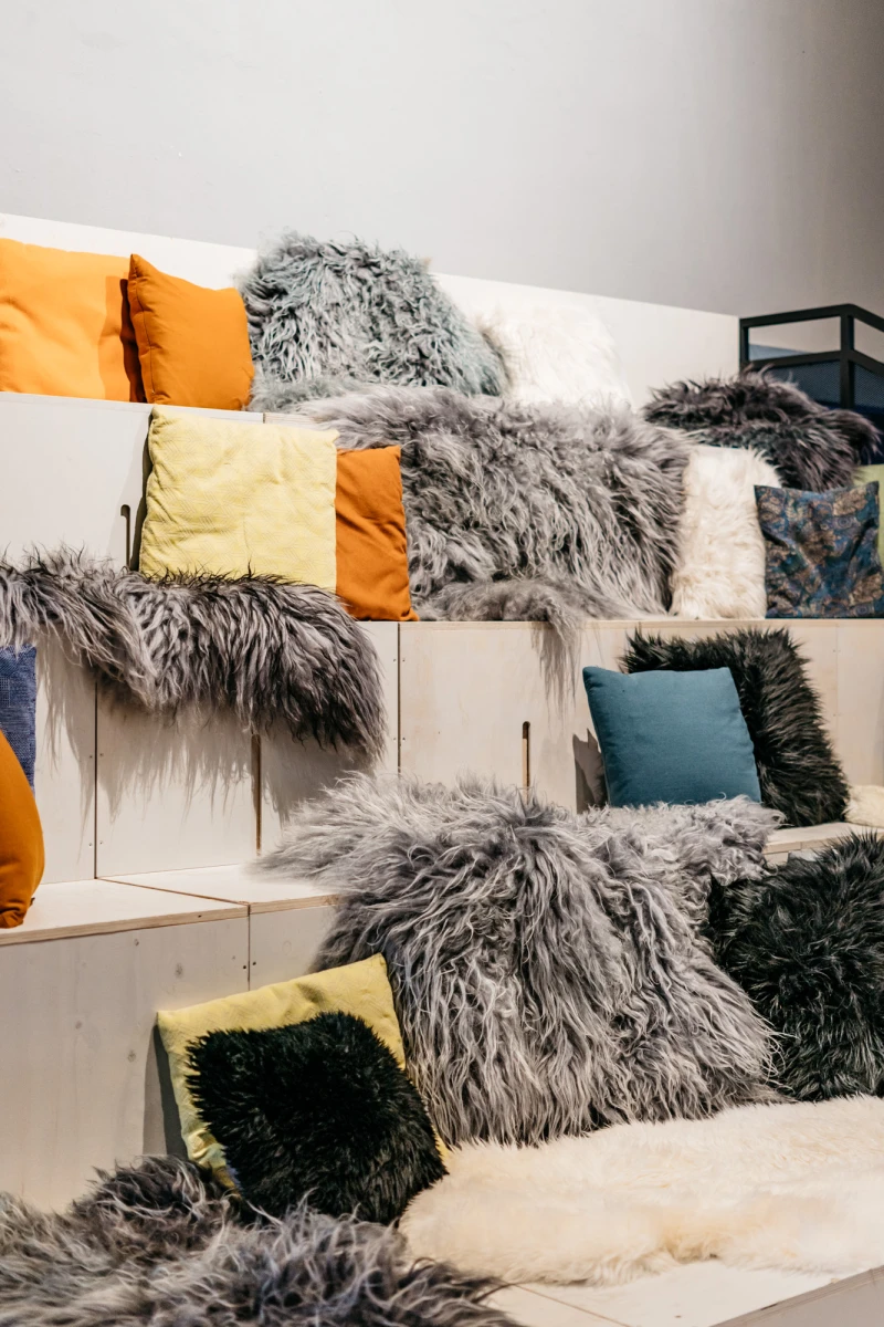 Gem&uuml;tliche Sitzgelegenheit mit mehreren Kissen in verschiedenen Farbt&ouml;nen wie Orange, Gelb und Petrol, bedeckt mit grauen, zotteligen Fell- und Pl&uuml;schkissen auf einer mehrschichtigen Sitzbank oder Treppe.