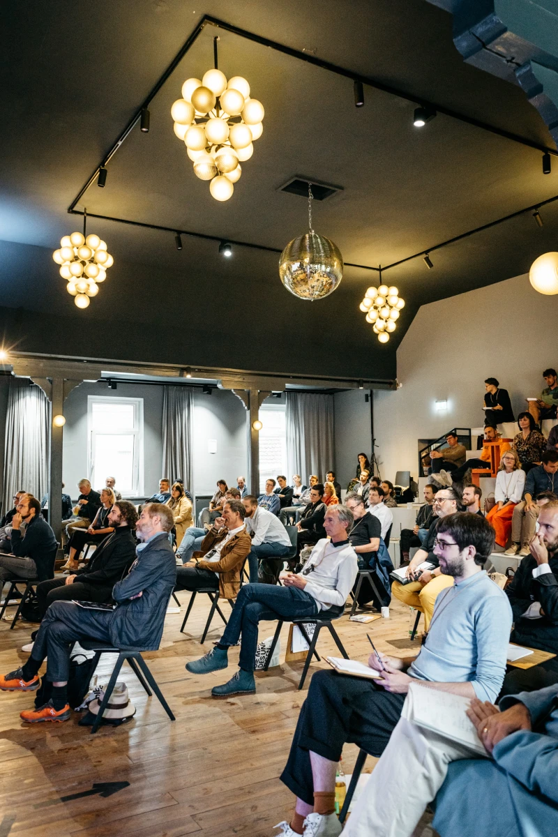 Teilnehmer einer Konferenz sitzen auf St&uuml;hlen in einem modern eingerichteten Raum mit auff&auml;lligen Deckenleuchten. Der Raum wirkt hell und einladend, mit Holzboden und gro&szlig;en Fenstern, w&auml;hrend die Anwesenden aufmerksam einer Pr&auml;sentation oder Diskussion folgen.