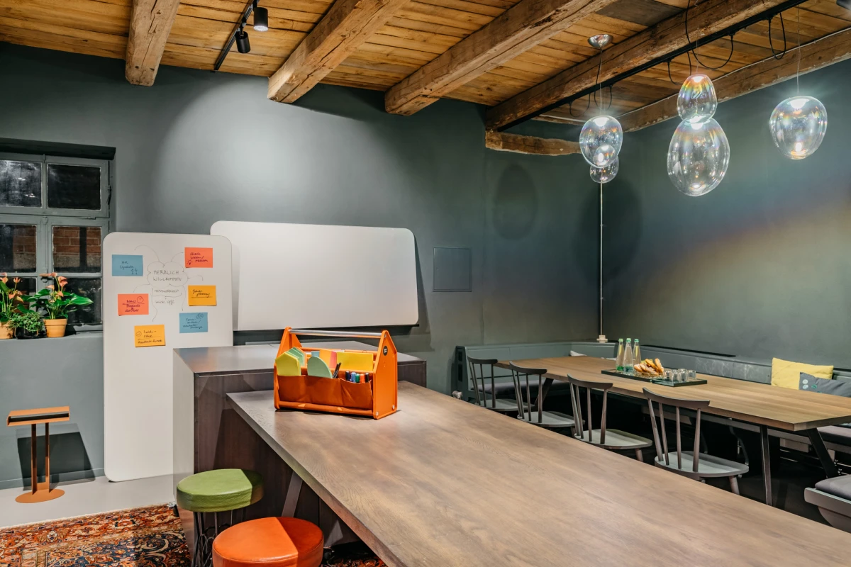 Moderne Teamk&uuml;che mit Holzdeckenbalken, grauen W&auml;nden und h&auml;ngenden Glaslampen. Ein langer Holztisch mit orangefarbener Werkzeugkiste und bunten Hockern, daneben eine Whiteboard-Wand mit Haftnotizen. Gro&szlig;e Fenster und eine gem&uuml;tliche Arbeitsatmosph&auml;re pr&auml;gen den Raum.