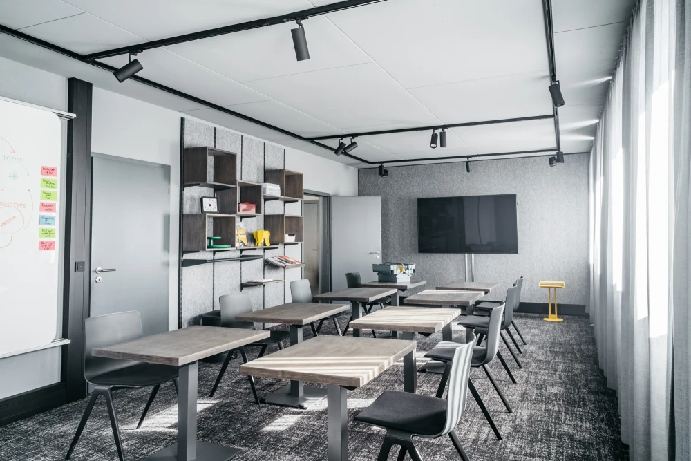 Moderner Besprechungsraum mit grauen W&auml;nden, mehreren Holztischen, schwarzen St&uuml;hlen und einer Wandregalkonstruktion. Schwarze Deckenspots und gro&szlig;e Fenster sorgen f&uuml;r helle, offene Atmosph&auml;re. Graue Teppichb&ouml;den und eine schwarze Wandtafel vervollst&auml;ndigen das minimalistische Design.