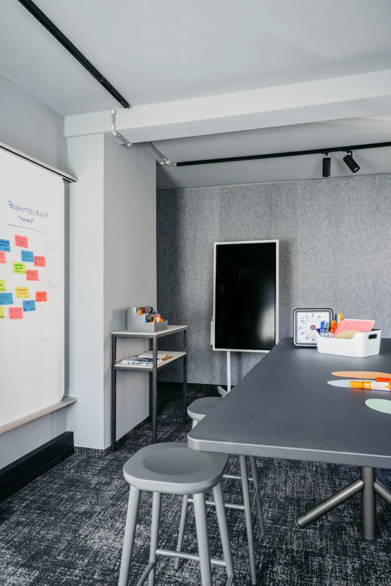 Moderner Besprechungsraum mit wei&szlig;er Wandtafel, Post-its, grauem Tisch, Hocker und schwarzer R&uuml;ckwand. Schwarze Deckenleuchten und Regale mit Utensilien vervollst&auml;ndigen das minimalistische B&uuml;roambiente.