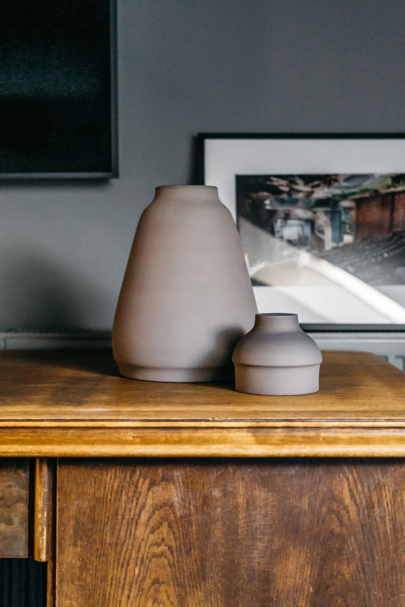 Zwei beige Vasen unterschiedlicher Gr&ouml;&szlig;e stehen auf einem Holzregal vor einem gerahmten Kunstdruck. Die Vasen haben eine matte, glatte Oberfl&auml;che und f&uuml;gen sich harmonisch in das minimalistisch-skandinavische Wohnambiente ein.