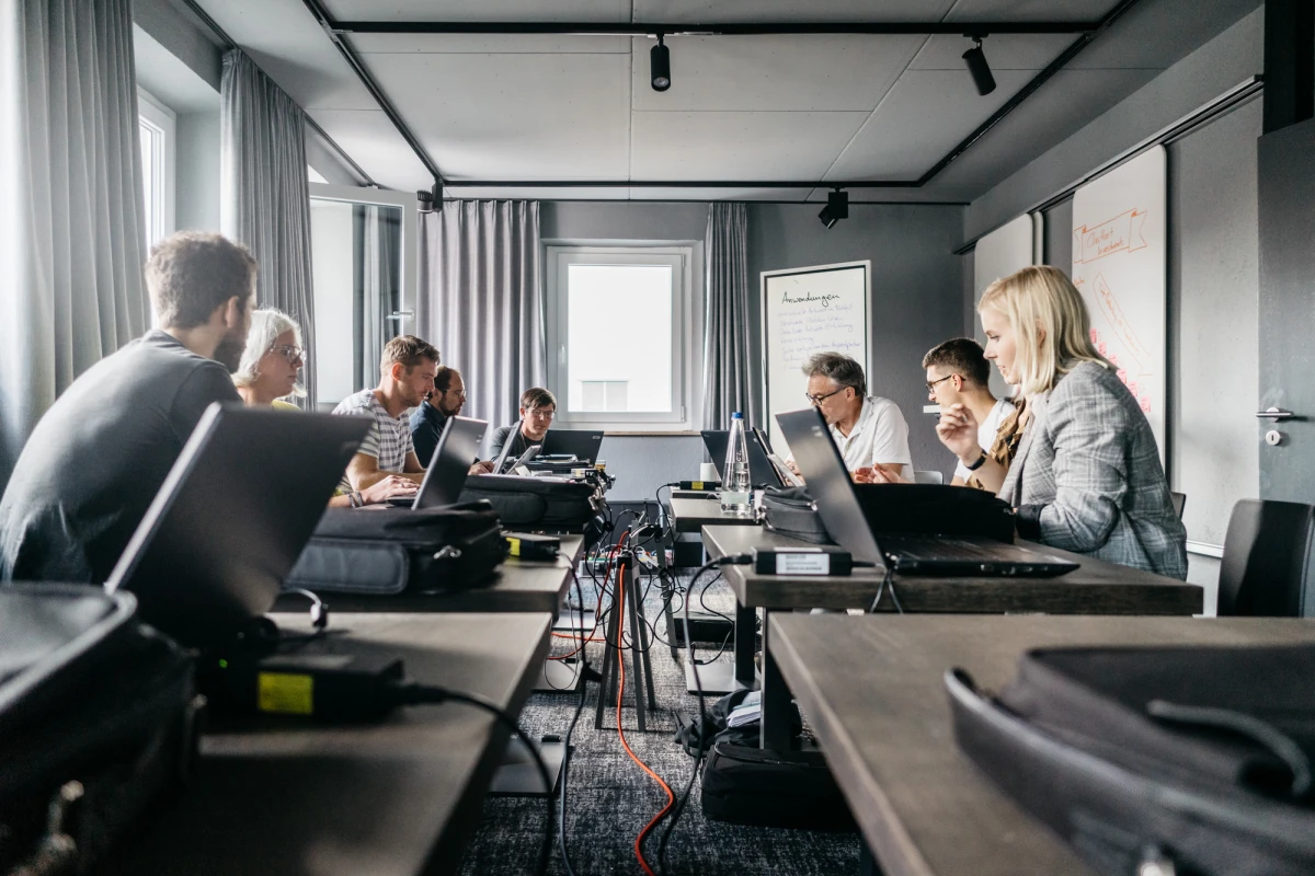 Mehrere Personen arbeiten konzentriert an Laptops in einem modernen Besprechungsraum mit grauen Vorh&auml;ngen und Fenstern. Die Teilnehmer sitzen an einem langen Tisch, auf dem Laptops, Kabel und technische Ger&auml;te verteilt sind. Die Atmosph&auml;re wirkt professionell und fokussiert.