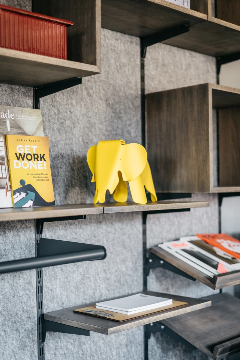 Graues B&uuml;cherregal mit gelber Elefantenfigur und B&uuml;chern, darunter ein gelbes Buch mit dem Titel "Work done". Die Regalbretter sind aus Holz und mit Filz ausgelegt, was eine gem&uuml;tliche und moderne Atmosph&auml;re schafft.