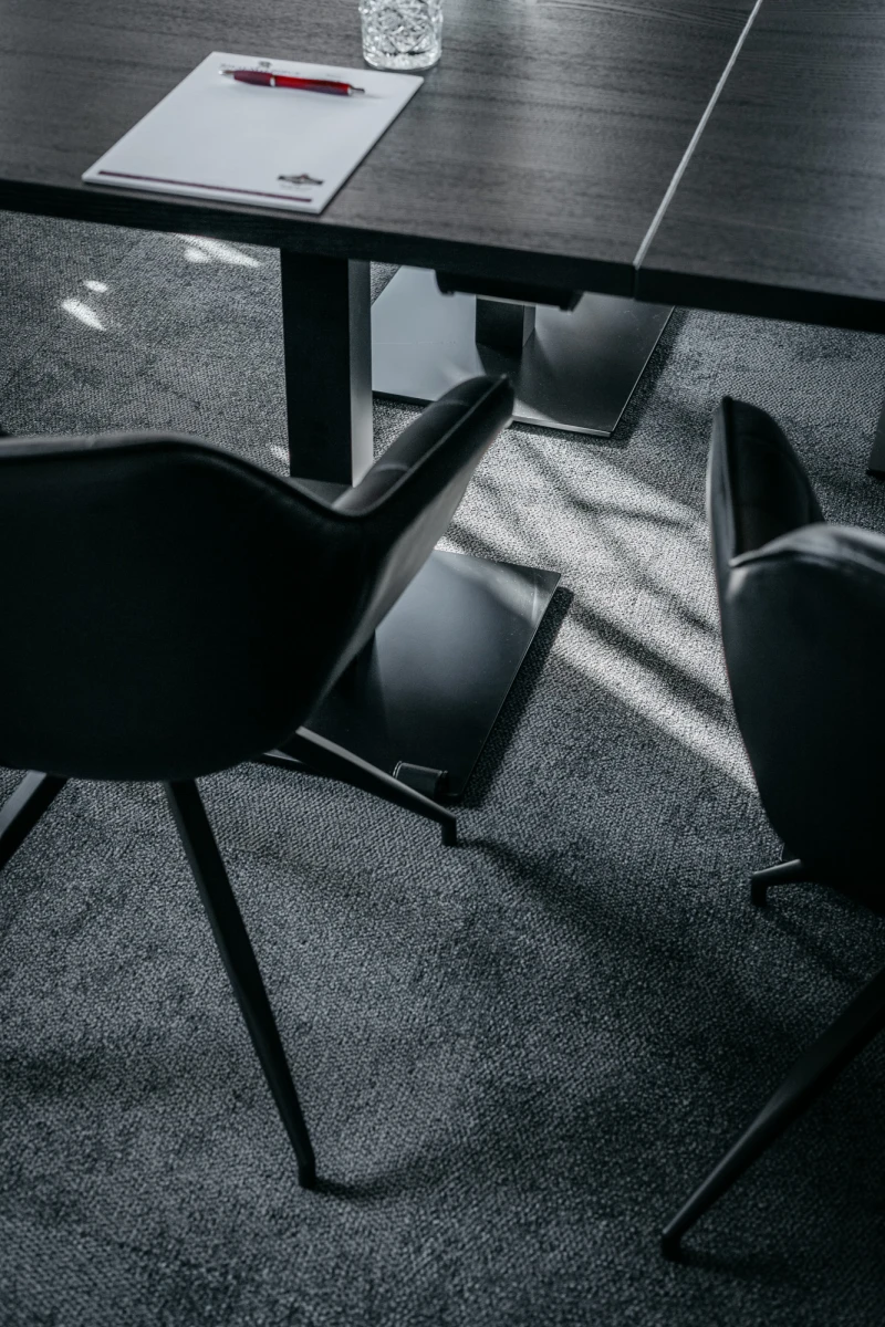 Moderner Konferenztisch mit Papier und Stift, umgeben von grauen Designerst&uuml;hlen auf einem dunkleren Teppichboden. Scharfe Lichtschatten fallen diagonal &uuml;ber die Szenerie und betonen die klare, geometrische Komposition des Raumes.