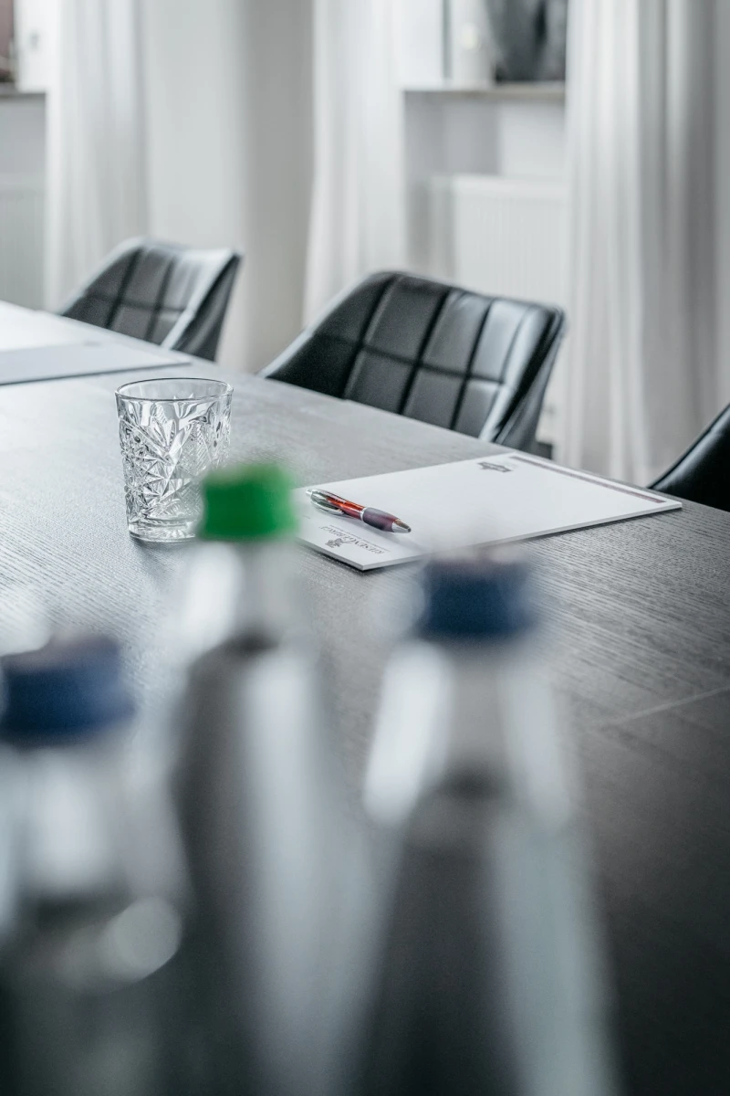 Holzerner Konferenztisch mit einem geschliffenen Wasserglas und einem Notizbuch, umgeben von schwarzen Lederst&uuml;hlen in einem hellen, minimalistisch eingerichteten Besprechungsraum.