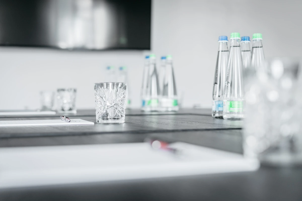 Transparente Wassergl&auml;ser und Glasflaschen stehen auf einem hellen Tisch oder Thekenbereich. Die Komposition wirkt minimalistisch und elegant, mit einem unscharfen Hintergrund in ged&auml;mpften Graut&ouml;nen.