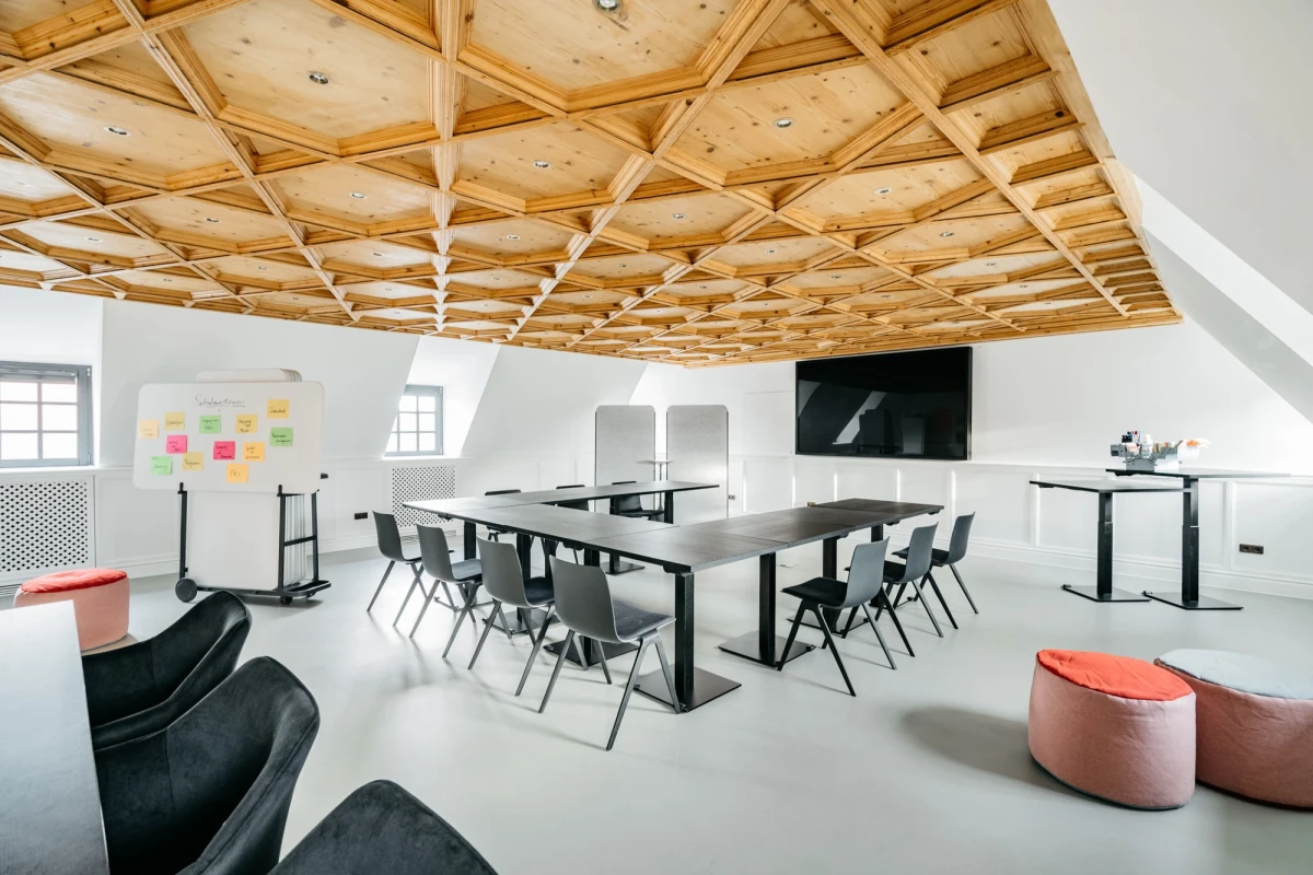 Moderner Konferenzraum mit Holzdeckenbalken und wei&szlig;en W&auml;nden, ausgestattet mit schwarzen Konferenzst&uuml;hlen, einem gro&szlig;en Meetingtisch, Flipchart und Fernsehbildschirm. Zwei rote Sitzpoufs und ein schwarzer Sessel vervollst&auml;ndigen die minimalistische Einrichtung.