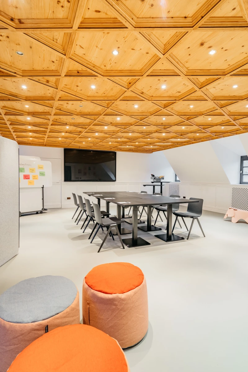 Moderner Konferenzraum mit Holzdecke und geometrischen Einbauleuchten, ausgestattet mit grauen B&uuml;rost&uuml;hlen um einen Konferenztisch, erg&auml;nzt durch orange und graue Sitzpoufs im Vordergrund und einer Pinnwand an der Wand.