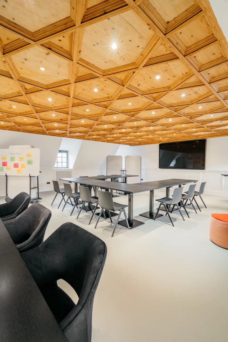 Moderner Konferenzraum mit holzget&auml;felter Decke und elegant designten M&ouml;beln. Graue Tische und St&uuml;hle stehen in U-Form, mit schwarzen Sesseln im Vordergrund und einer wei&szlig;en Wand mit bunten Haftnotizen. Warmes Licht und gro&szlig;e Fenster sorgen f&uuml;r eine einladende Arbeitsatmosph&auml;re.
