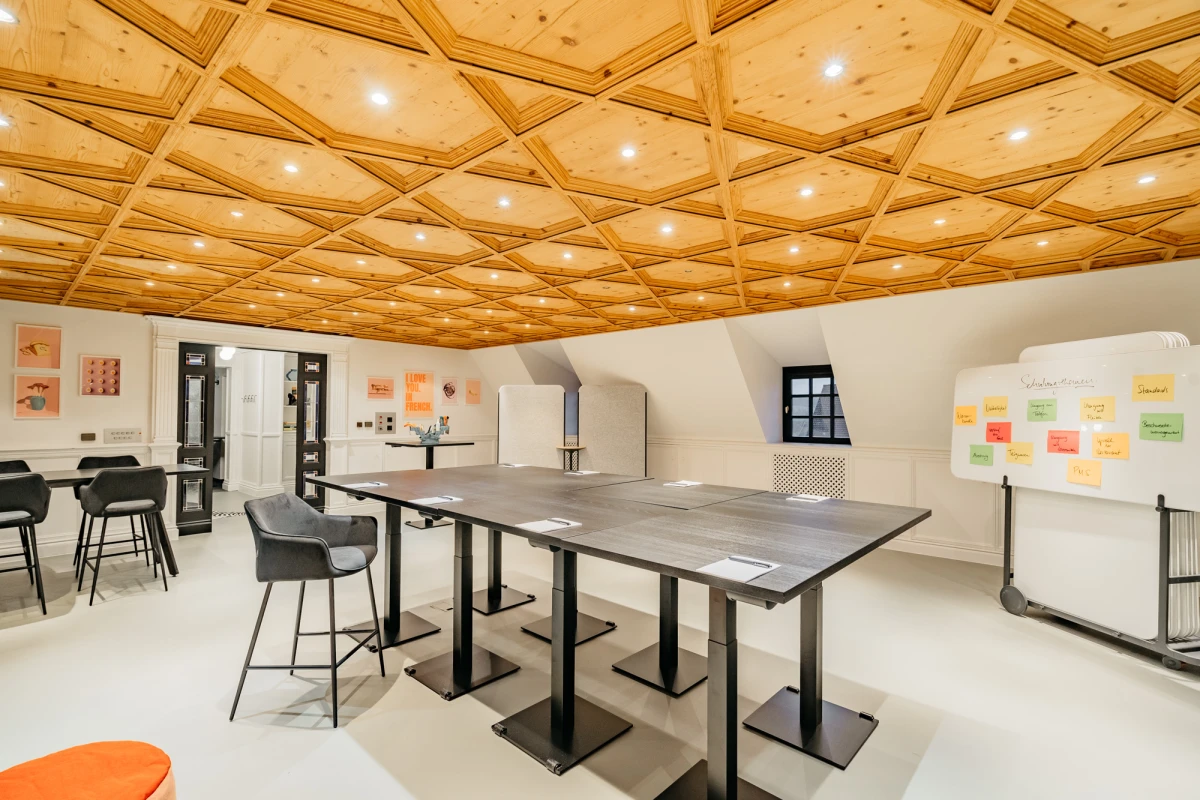 Moderner Besprechungsraum mit elegantem Holzdeckendesign, grauen Arbeitstischen und schwarzen St&uuml;hlen. Helle, offene Atmosph&auml;re mit eingebauten Deckenleuchten und einer Pinnwand an der Wand. Professioneller Arbeitsbereich mit nat&uuml;rlichem Licht und zeitgem&auml;&szlig;em Mobiliar.