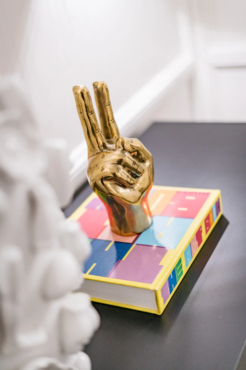 Goldene Hand-Skulptur mit Victory-Zeichen, die auf einem bunten, mehrfarbigen Buch oder Kasten mit gelben Kanten sitzt. Die Skulptur ist auf einer dunklen Oberfl&auml;che vor einem hellen Hintergrund platziert.