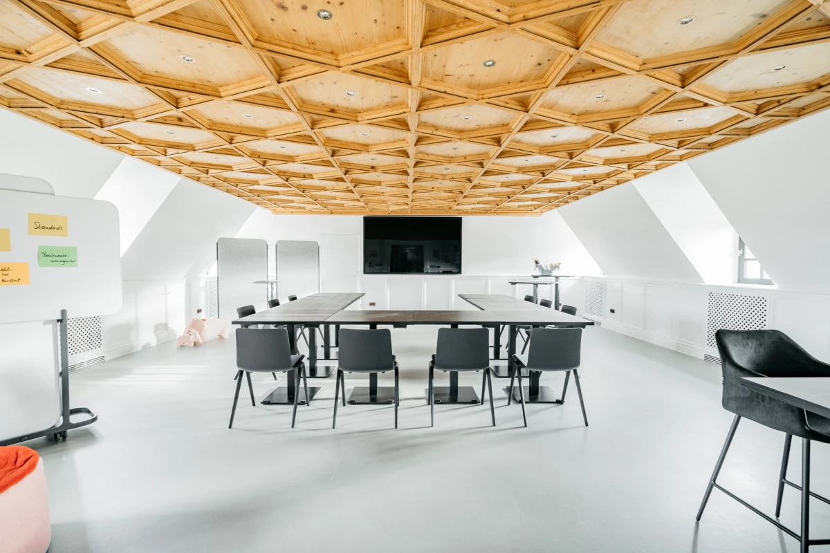 Moderner Besprechungsraum mit geometrischer Holzdecke, wei&szlig;en W&auml;nden und schwarzen St&uuml;hlen um einen rechteckigen Tisch. Ein schwarzer Bildschirm h&auml;ngt an der Wand, und der Raum wirkt hell und minimalistisch gestaltet.