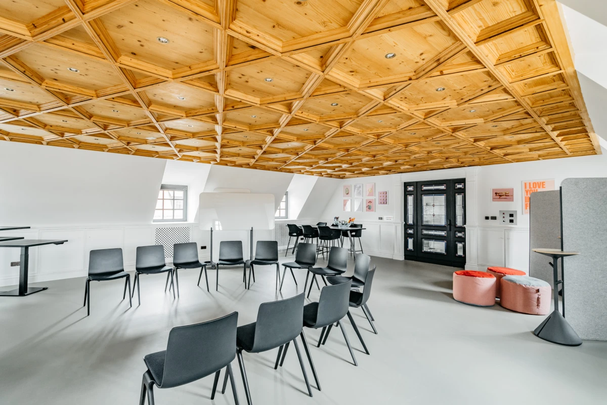 Heller Seminarraum mit holzget&auml;felter Decke und modernen grauen St&uuml;hlen, die in Reihen angeordnet sind. Zwei Fenster und eine schwarze T&uuml;r an der Wand, daneben rosa Sitzpuffs und technische Ger&auml;te.